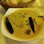 A bowl of ema datshi: a pale cheese sauce with two whole dried red chillies and pieces of meat, and a basket of steamed buns behind it