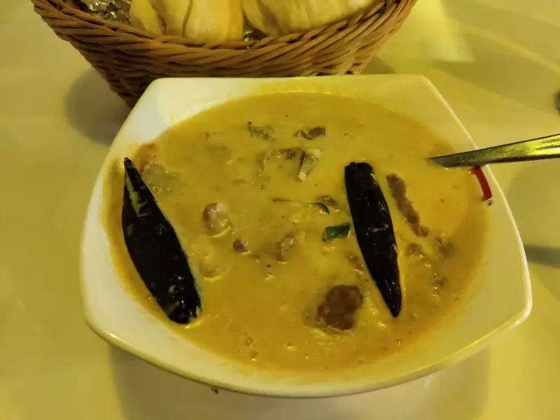 A bowl of ema datshi: a pale cheese sauce with two whole dried red chillies and pieces of meat, and a basket of steamed buns behind it